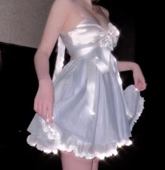 a woman wearing a white dress with lace trim and a bow at the neckline is standing and looking away. the dress has a blue tint and is gathered around her waist. she is standing in front of a dark background.