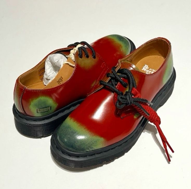 a closeup shot of a pair of dr. martens shoes. the shoes are a unique tiedye design with a red and green color scheme. they are laceup oxfords with a chunky sole. the shoes are stylish and trendy, and would be a great addition to any wardrobe.