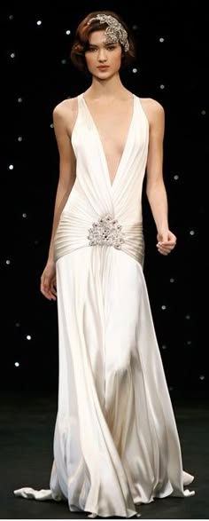 a beautiful woman wearing a white, elegant gown, posing for a fashion shoot. she has a vintage, flapperinspired headpiece and jewelry. her dress is a classic style with a deep vneck and a flowing skirt. the model's hair is styled in a chic updo, and her makeup is flawless. she is radiating confidence and glamour.