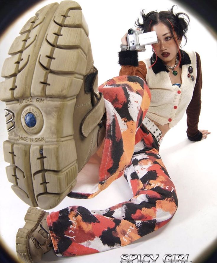 a young woman wearing red, black, and white splatter paint pants poses while holding a camera. she has on a pair of oversized brown boots.