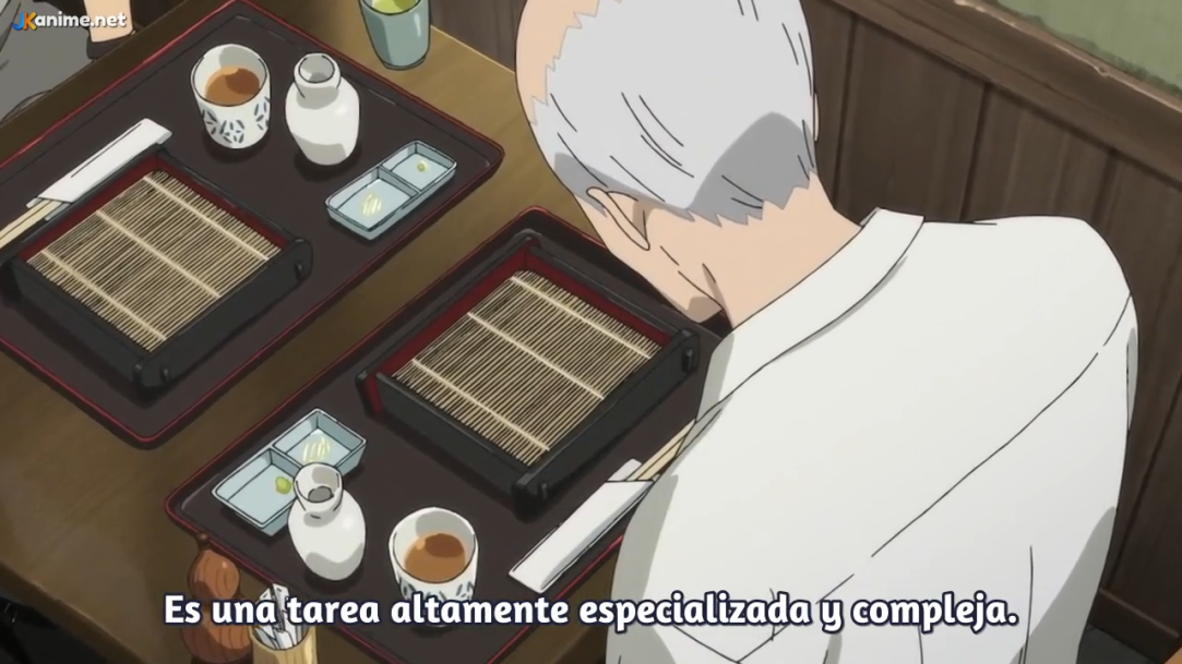 a behind the shoulder shot of a man with grey hair wearing a white shirt seated at a table. on the table are two place settings, each with a black tray of food, two white dishes, a white cup, and a white pitcher. the wall in the background is brown with vertical wood paneling.  the text "es una tarea altamente especializada y compleja." is at the bottom of the image.
