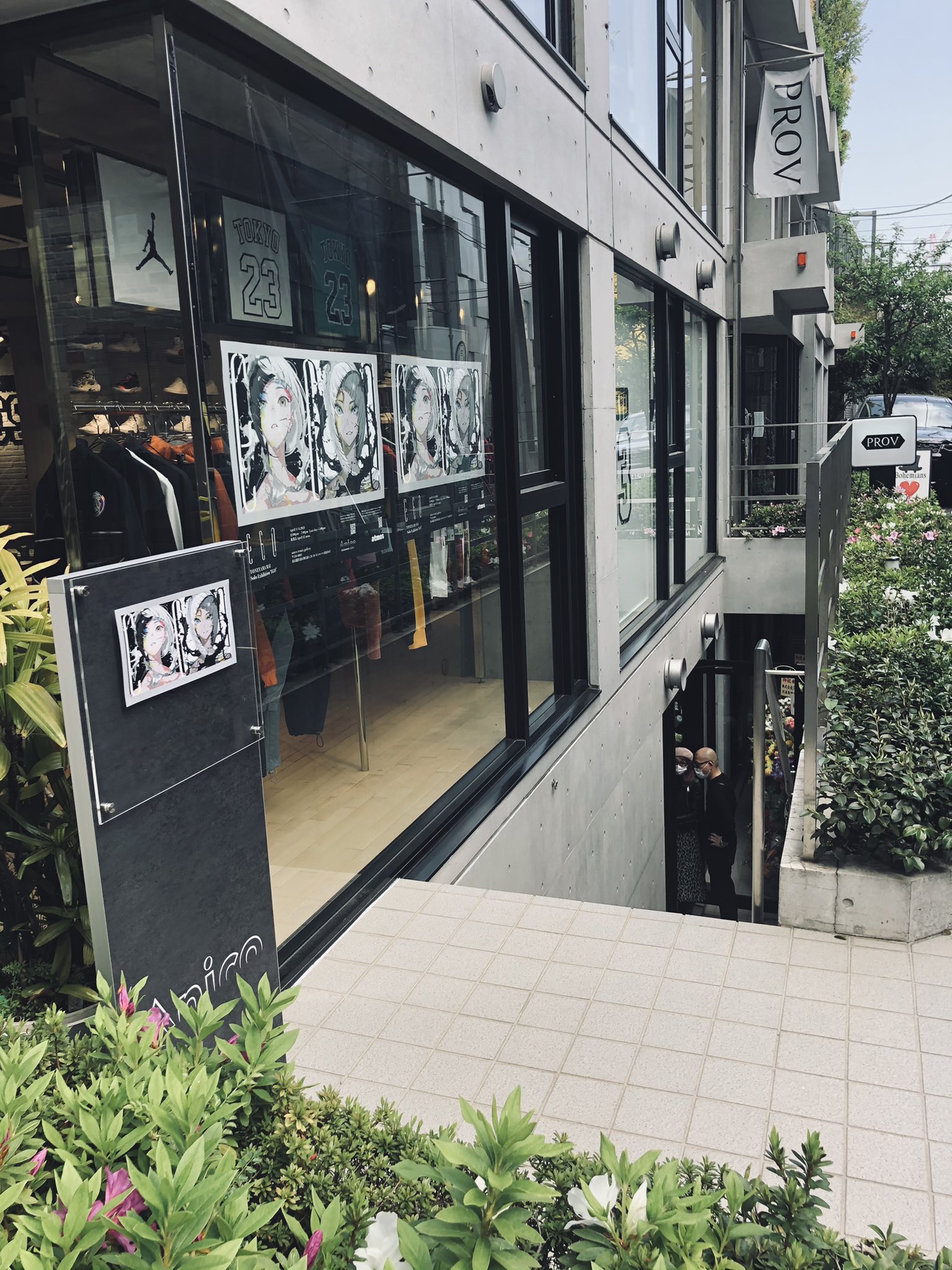 the image shows the exterior of a streetwear store, likely located in japan. the storefront is modern and minimalist, with large glass windows displaying clothing and sneakers. there are two people entering the store, which is located below street level. the overall vibe is trendy and urban.