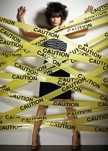 a woman in a striped dress is wrapped in yellow caution tape.  the tape is crisscrossed around her body, leaving her bound and unable to move.  she is wearing high heels and has a concerned expression on her face.