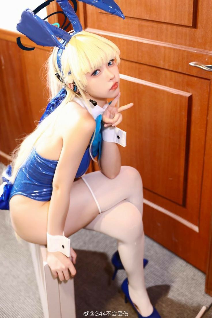 a woman in a blue bunny costume with blonde hair, white tights, and blue high heels sits on a lightcolored surface indoors. she's holding up a peace sign. a light brown door is behind her.