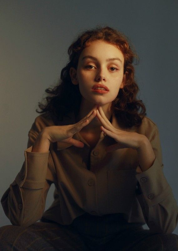 a portrait of a young woman with curly brown hair sitting in a studio with her hands clasped together under her chin. she is wearing a brown shirt and looking directly at the camera.