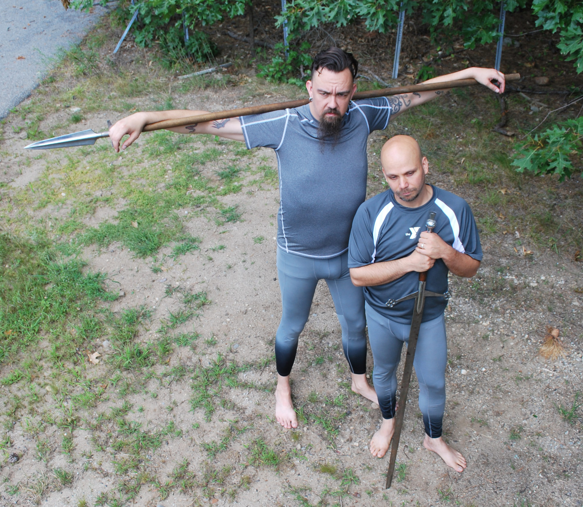 two athletic men stand outside in the dirt, grass and trees. one man holds a spear over his shoulder and smiles, while the other man holds a sword and looks directly at the camera. both men are wearing grey and black athletic gear.
