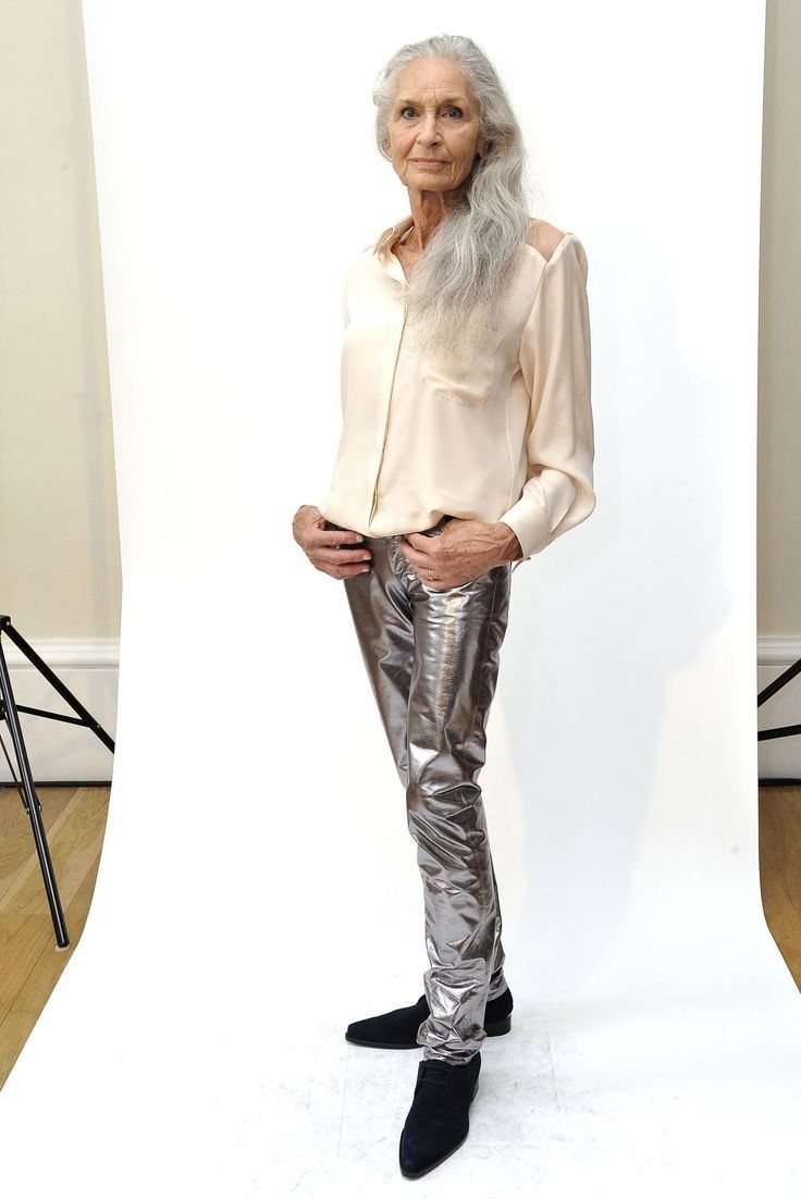 a fulllength studio shot of an elderly woman with long silver hair. she is wearing a creamcolored, loosefitting shirt and silver metallic pants. she is posing with her hands in her pockets, and her expression is serious. the background is plain white.  she's wearing black boots. the overall style is elegant and stylish.
