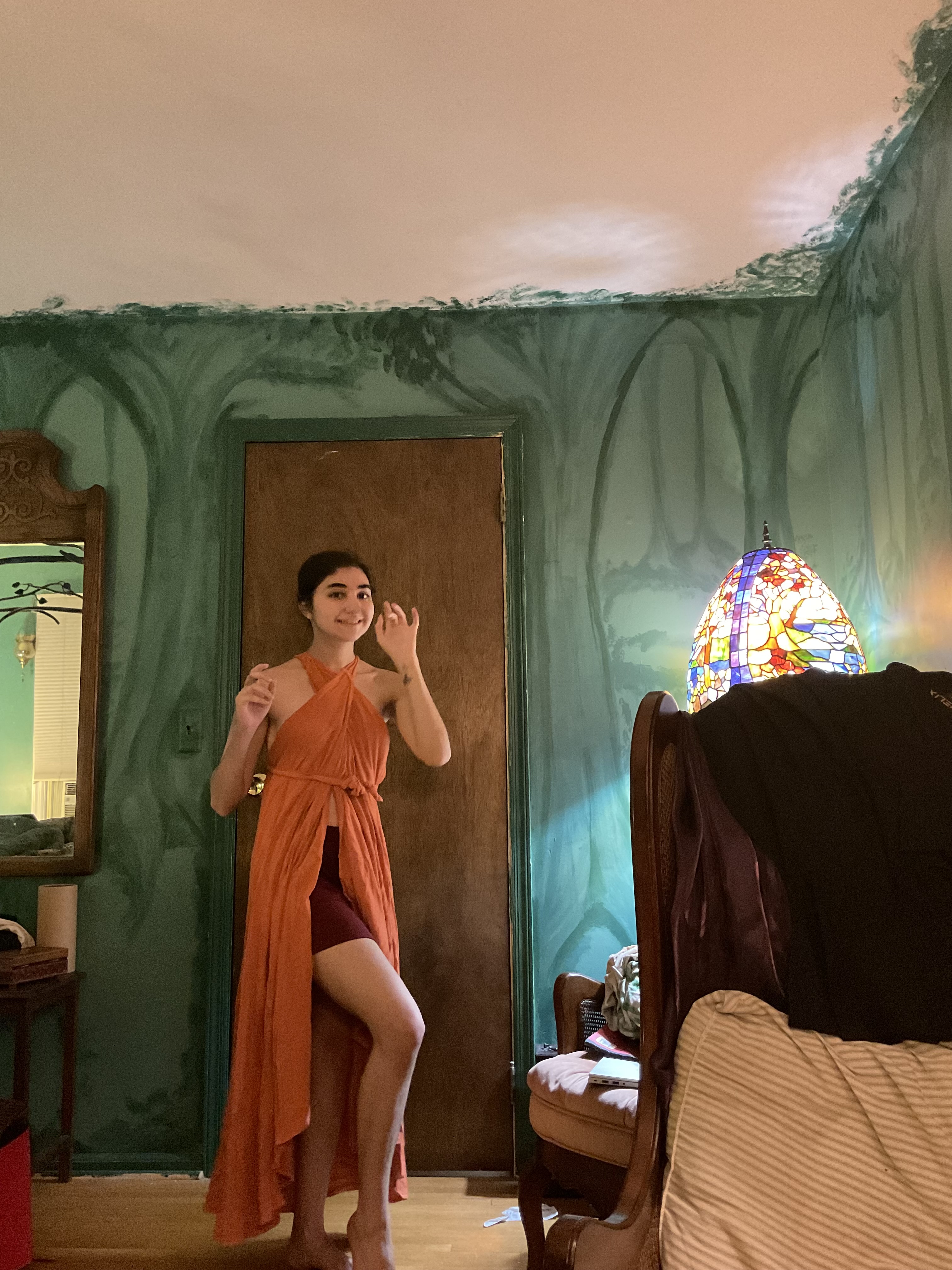 a young woman is standing in a bedroom with a uniquely painted green wall featuring treelike designs. she is wearing a long, orange wrap dress that is highlow in style, and is posing slightly towards the camera. the room is furnished with a chair and a mirror, and a stained glass lamp is visible in the background. the overall setting and style are somewhat bohemian and artistic.
