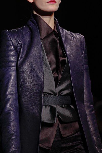 a woman in a dark purple leather jacket over a black satin shirt, with a small black bow at the neck, is looking off to the side. her black belt is visible just below the shirt.  the background is a dark, outoffocus color, and the woman's expression is serious.