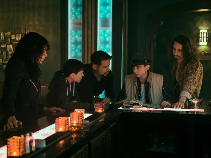 five people are gathered around a dimly lit bar, seemingly engaged in an intense conversation. the warm, spotlit lighting creates a sense of mystery and intrigue. the group is comprised of two women, two men, and a teenage boy, suggesting a possible family dynamic.  a book lies open on the bar, potentially holding information relevant to their discussion. the overall atmosphere hints at a clandestine meeting or a moment of suspense.