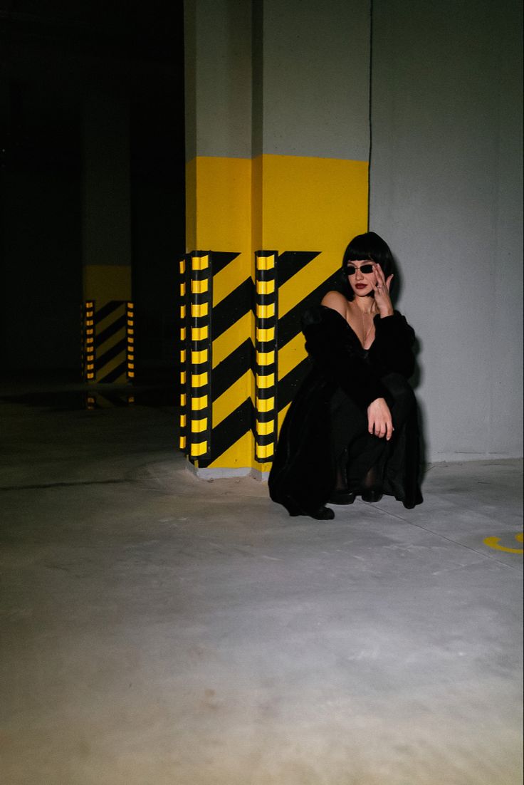 a woman in a black coat and sunglasses sits on the floor of a parking garage. she is leaning against a pillar with yellow and black stripes. the floor is concrete and the walls are gray.