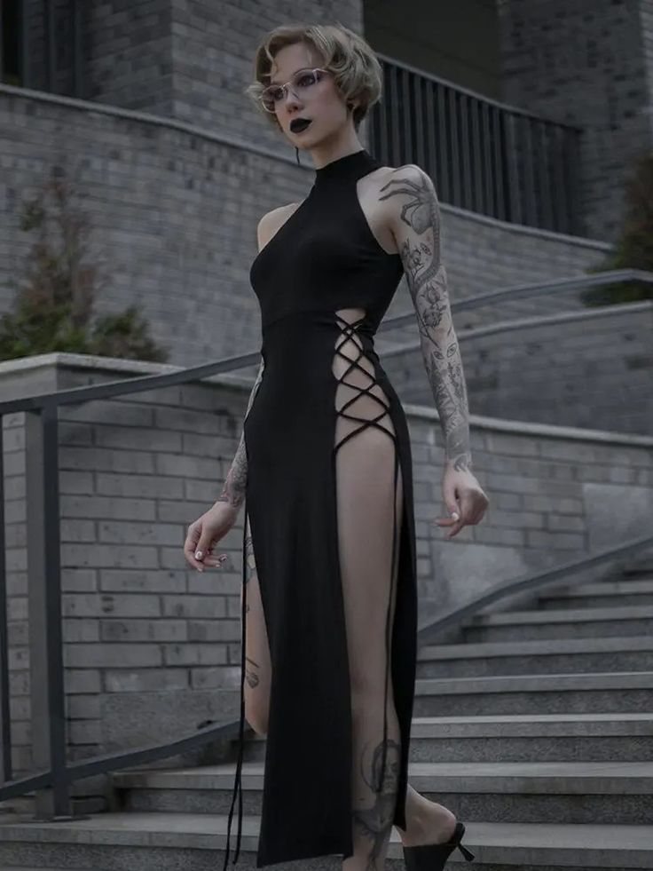 a stylish woman in a black, long, sideslit dress, with lace detailing on the slit, stands on stairs with a confident pose. she's wearing high black heels and her left leg is clearly visible due to the high slit. she has several tattoos visible on her arms and leg, adding an edgy, stylish touch to the look.