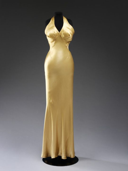 a gold colored halter style gown is displayed on a mannequin. the gown is made of satin or silk and has a vintage look to it.