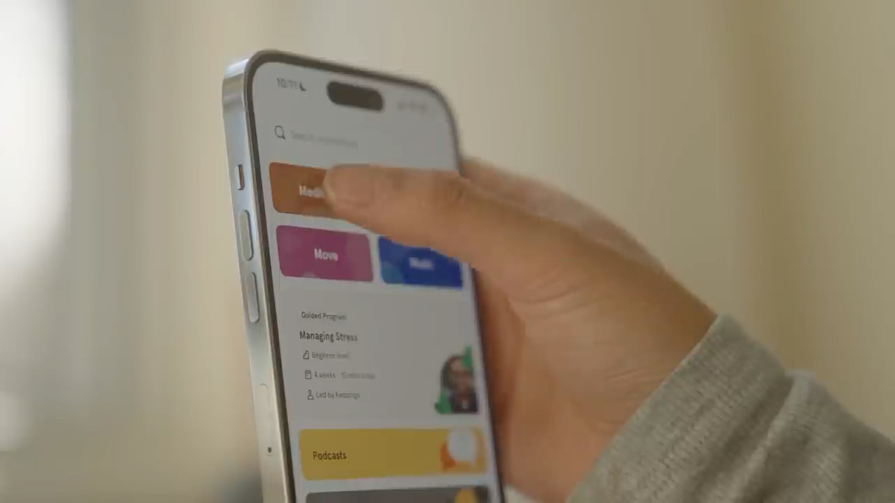 a closeup shot of a person's hand as they use their index finger to scroll through a wellness or mental health app on their smartphone. the app's interface is visible, displaying options like "managing stress", "move", and other categories like "podcasts". the background is blurred, indicating an indoor setting, and the lighting is soft and bright.  the phone has a unique, rounded silver edge.