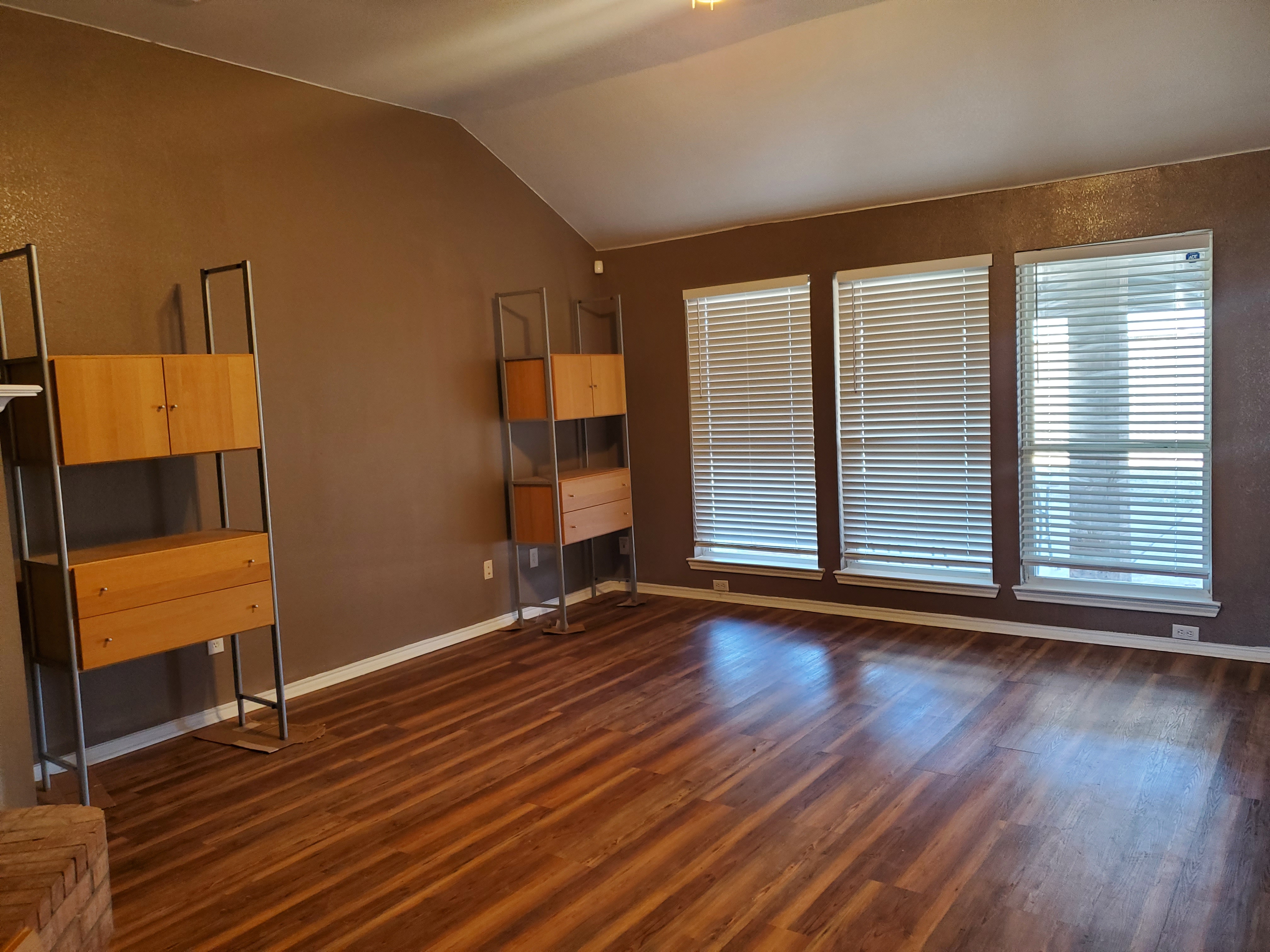 a living room with brown walls, wood floors, three windows with blinds, two shelving units, and a vaulted ceiling.