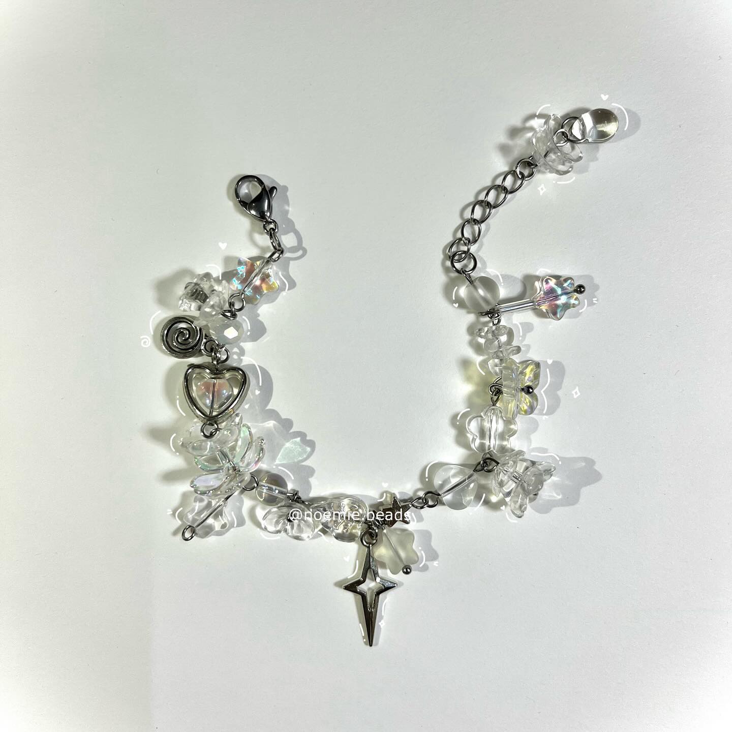 a handmade bracelet featuring iridescent and clear beads, flower beads, a star charm, a heart charm, and a spiral charm. the bracelet has a silver chain and a lobster clasp. it is displayed on a white background. the bracelet is designed and made by doemie beads.