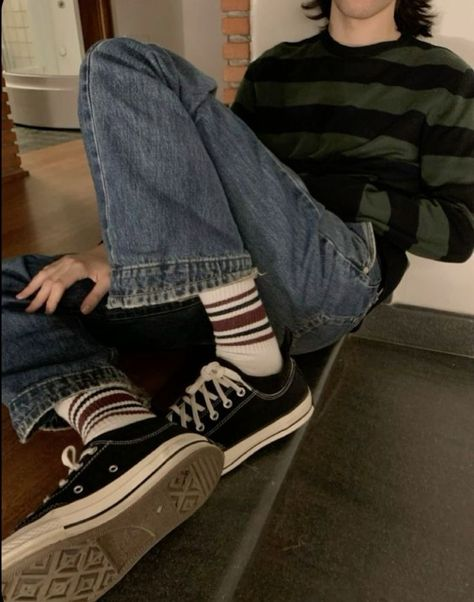a young man wearing a striped sweater, black converse shoes with striped socks, and blue jeans. he's leaning against a wall and looking at the camera. the image is casual and stylish, with a focus on the man's footwear and clothing.