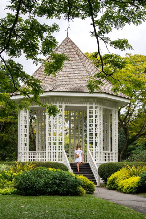 a young woman in a white dress walks down the stairs of a white gazebo in a lush green garden setting.  the gazebo is octagonal with a shingled roof and intricate white latticework.  the garden is filled with various plants and trees, creating a vibrant and peaceful atmosphere.  the image suggests a location like the singapore botanic gardens.