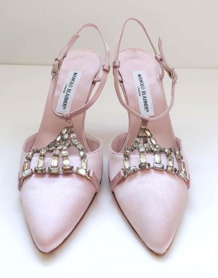 the image shows a pair of manolo blahnik heels in a light pink satin.  the shoes are pointedtoe, tstrap style and feature a delicate embellishment of clear crystals across the vamp.  the manolo blahnik label is visible on the inner sole of each shoe. the shoes are elegantly positioned against a light background. they appear to be new or in excellent condition.