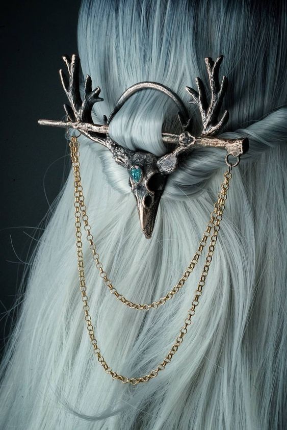 a close up image of a silver skull antler hair piece  with a blue jewel.  the hair piece is on a model with grey hair.  the piece is adorned with a gold chain, that drapes from the bottom of the piece.