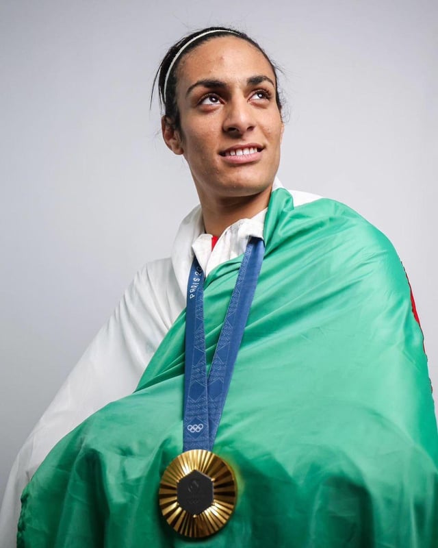 an algerian or moroccan female athlete, beaming with pride, draped in her national flag. she wears a white athletic top and her hair is covered with a black headband. the focus is on her joyful expression as she holds a gold medal, likely earned at the olympic or paralympic games.