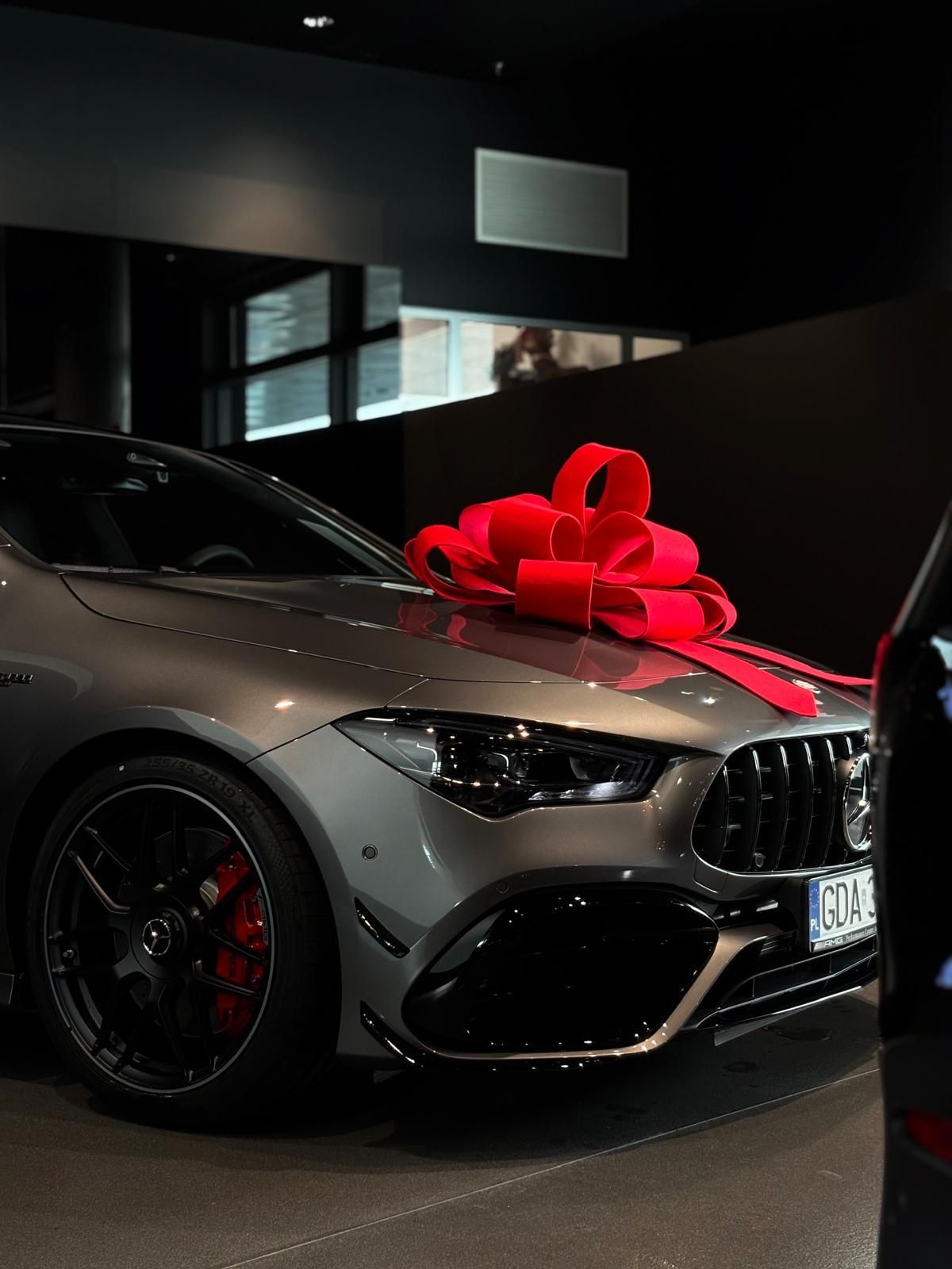 a grey mercedesbenz with a large red bow on the hood is parked in a car dealership. the car has black rims and red brake calipers. another car is partially visible on the right side of the frame. the background is dark, with a window and a dark wall.