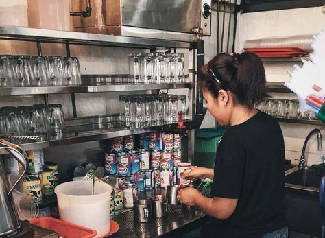 a woman is working behind the counter of a busy cafe. she is making coffee or tea, using various equipment such as pitchers, cups, and cans of condensed milk.  the cafe's kitchen is wellstocked, with shelves filled with glasses and other supplies. the scene is busy and suggests a bustling atmosphere of a restaurant or eatery.  the woman is focused on her task and appears to be skilled in her work.
