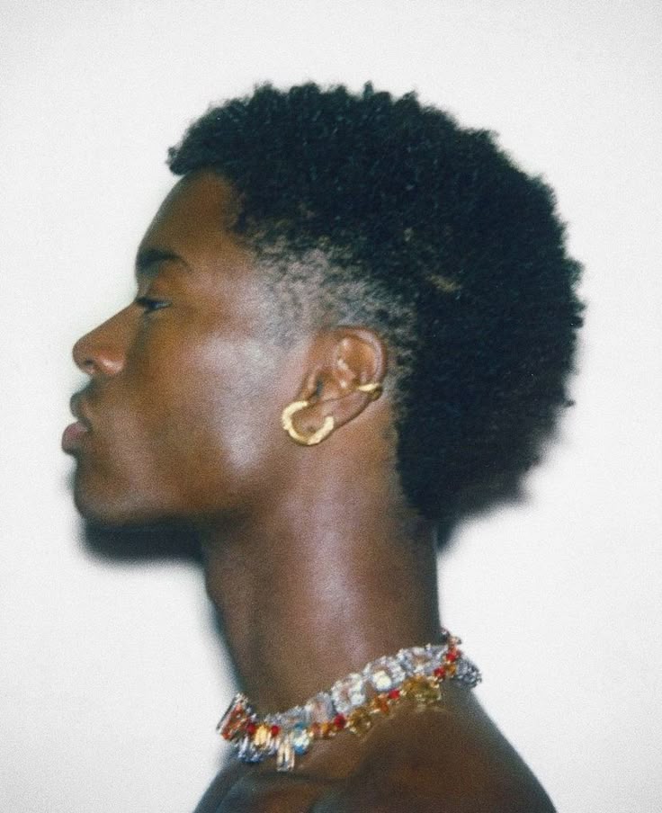 a closeup, sideview portrait of a darkskinned man wearing jewelry. he has short, dark hair with a fade haircut. he is wearing a gold earring and a colorful jeweled necklace.