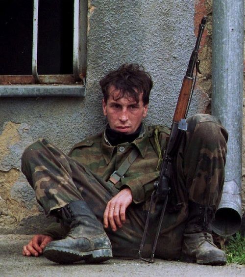 a young soldier in camo fatigues sits near a building, clutching a rifle. he looks tired and wary, with a determined expression on his face.  he is likely in a warzone and awaiting the next move.