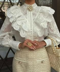 a woman wearing a white lace blouse with frilly details and pearl buttons. she is also wearing a pearl necklace and a gold ring. the blouse is paired with a white tweed skirt. she is holding a white quilted handbag with a gold chain strap.