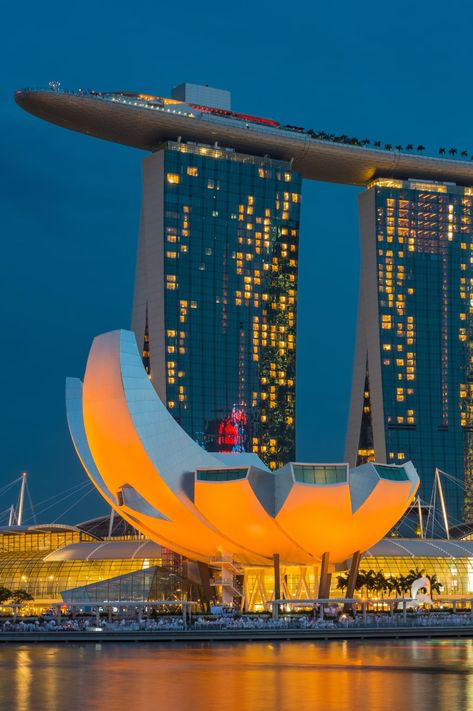 the image showcases a stunning nighttime view of the marina bay sands integrated resort in singapore.  the iconic hotel, with its distinctive shiplike structure perched atop three towers, is brightly lit, contrasting beautifully against the darkening twilight sky.  in the foreground, the artscience museum, with its unique flowerlike design, is also illuminated in a warm orange glow. the calm waters of marina bay reflect the city lights, creating a mesmerizing scene. the overall impression is one of modern architecture, luxury, and vibrant urban life.