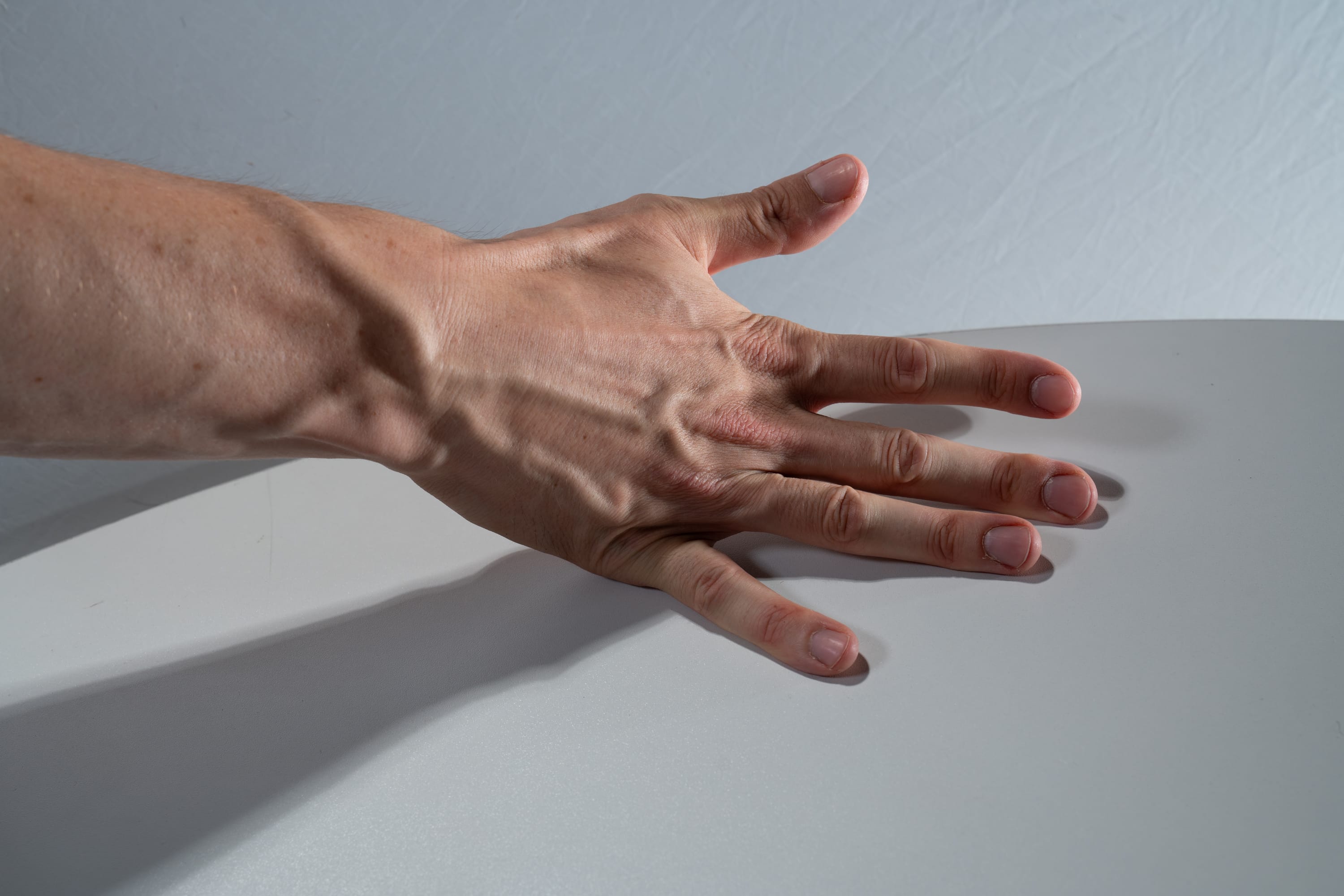a closeup view of a man's hand resting on a white surface. the hand is extended with fingers slightly apart, showcasing prominent veins and tendons on the skin. the lighting is bright, highlighting the details and textures of the hand.  the image focuses on the hand's anatomy and the interplay of light and shadow on its surface.