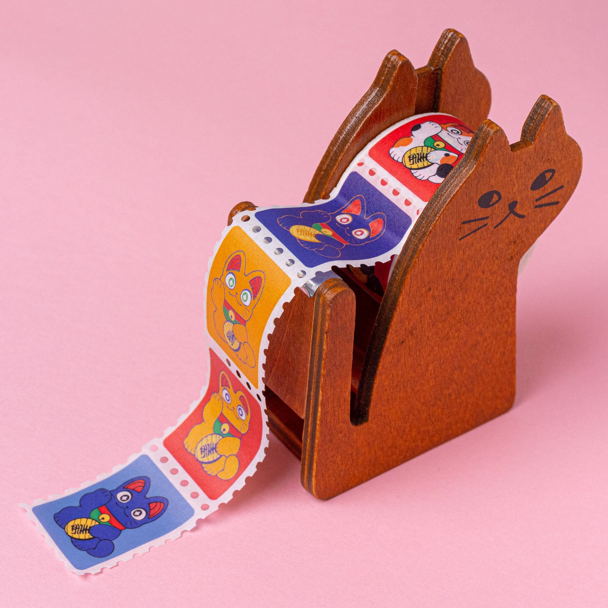 a closeup shot of a cute wooden catshaped sticker dispenser on a pink background. the dispenser is dispensing a roll of stickers featuring various illustrated manekineko (lucky cat) designs in different colors. the stickers are designed in a stamplike format. the overall aesthetic is kawaii and playful, showcasing japaneseinspired imagery.