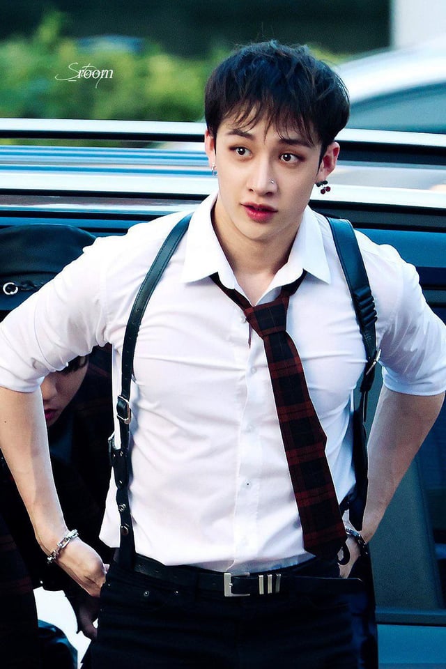 a closeup shot of bang chan, a member of the south korean kpop group stray kids. he's wearing a crisp white shirt with a red and black plaid tie, black suspenders, and dark pants.  he has dark hair styled neatly and is wearing an earring.  his expression is serious and intense. the background is blurred, with a dark car partially visible. the overall impression is one of stylish and sophisticated cool.