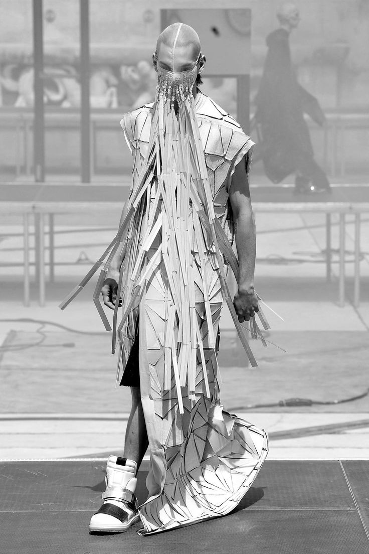 a monochromatic photograph captures a model on a runway showcasing an avantgarde, futuristic fashion piece. the model is wearing a geometric outfit adorned with strips and a mask that obscures the face. the ensemble is paired with hightop sneakers. the background features the runway setting and additional figures, blurred to emphasize the primary subject.