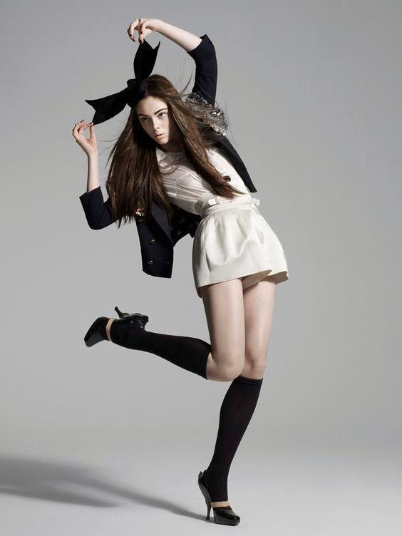 a slender, paleskinned model with long brown hair poses against a light grey background in a studio setting.  she wears a white silk romper, black jacket, black ankle socks and black heels.  a large black bow adorns her hair. she is striking a dramatic pose with one leg extended, her arms above her head and her gaze at the camera.