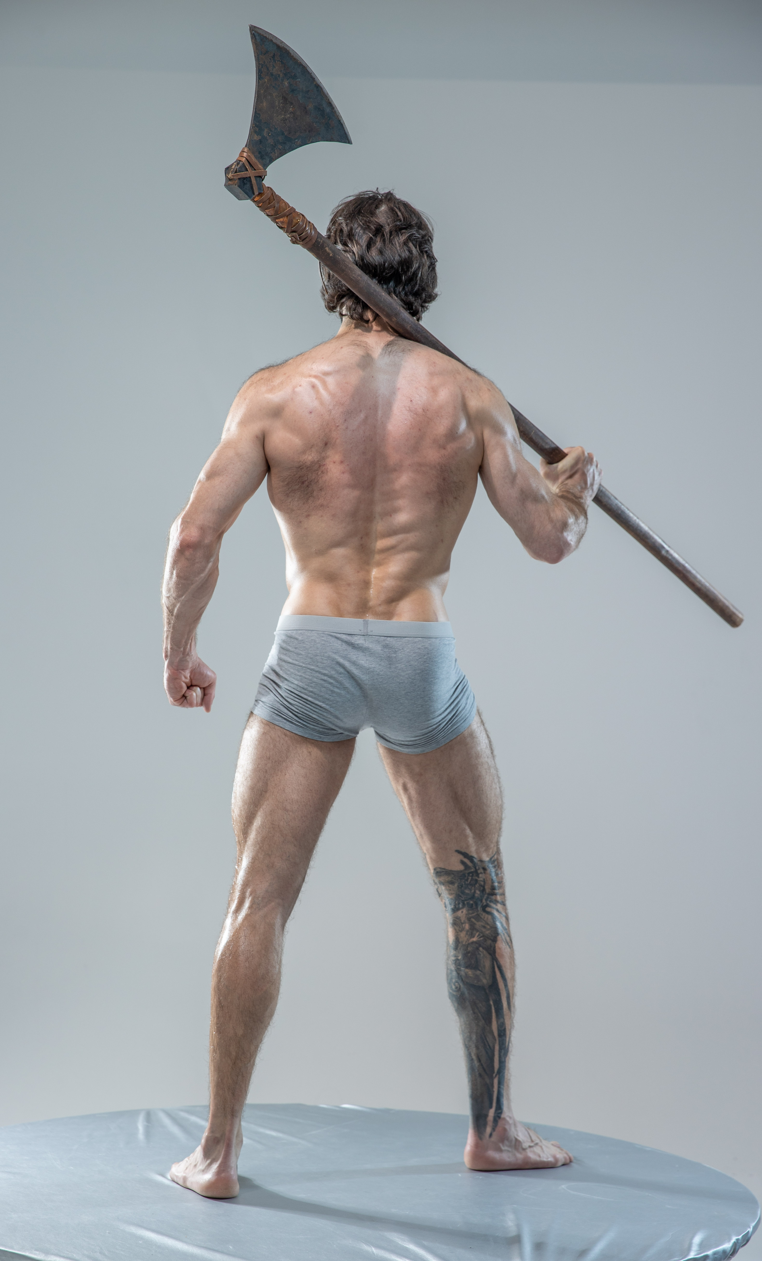 a muscular man with a large tattoo on his left calf stands with his back to the viewer on a small round platform. he holds a large, worn axe over his right shoulder. he is wearing only grey underwear. the background is a solid light gray. the lighting is soft and diffused.