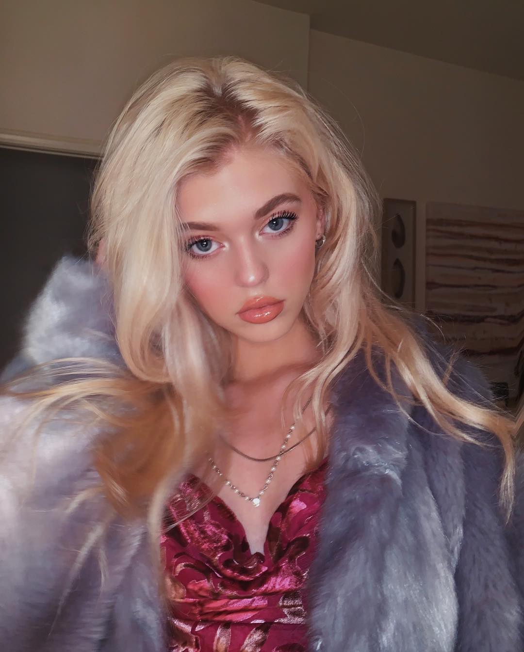 a portrait of loren gray, a young woman with blonde hair and blue eyes, wearing a pink floral dress and a grey faux fur coat. she is also wearing a delicate necklace. the photo is taken indoors and highlights her fair skin and glossy lips.