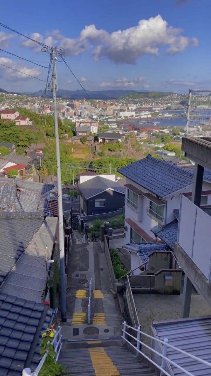 a vertical, eyelevel shot looks down a steep set of concrete stairs painted with yellow stripes, leading through a residential area towards a distant city skyline under a partly cloudy sky. houses and buildings flank the stairs on both sides, with visible rooftops and varying architectural styles. a utility pole with wires runs along the left side of the stairs.