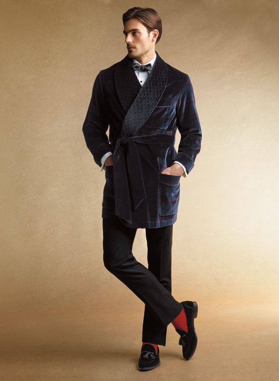 the image showcases a male model elegantly dressed in a dark blue velvet smoking jacket or robe.  the robe is a shawl collar design with a quilted inner lining visible at the collar. he is wearing black dress trousers, black tassel loafers, and vibrant red socks, adding a touch of unexpected color.  his shirt and bow tie are formal, suggesting a luxurious and sophisticated style. the background is a simple, muted beige, allowing the model and his attire to be the focal point. the overall aesthetic suggests a classic, perhaps vintageinspired, style with highend quality fabrics and tailoring.