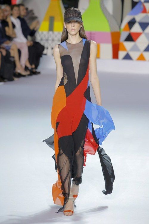 a model walks down a runway wearing a colorful, geometricpatterned dress. she is sporting a black cap and her hair is pulled back from her face. the background is brightly colored, creating a vibrant contrast with the model's attire. her pose exudes confidence and grace, showcasing the intricate details of the designer's creation.