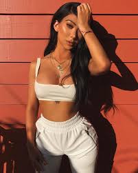 the image shows a young, attractive woman with long, dark brunette hair. she's wearing a white crop top and matching white sweatpants. she's accessorized with several gold necklaces and a bracelet. the woman is posing confidently against a reddishorange wall, with her hand gently resting on her hair.  the lighting suggests an outdoor setting, likely during the day. her tan skin is visible, and the overall style is trendy and fashionable. the background is predominantly a vibrant reddishorange color with distinct horizontal lines that provide a backdrop to her figure.  there's a visible shadow cast on the wall to the right of the model, accentuating the light playing on her face and body.