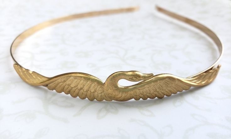 a delicate gold headband with a swan design. the swan is in flight, with its wings outstretched. the headband is simple and elegant, and would be perfect for a special occasion.