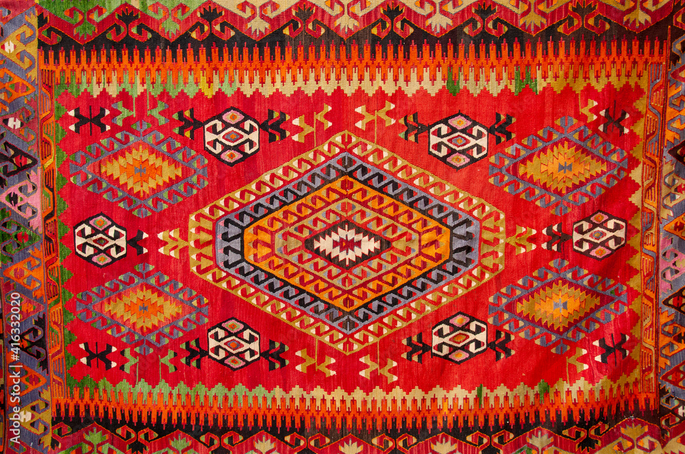 the image shows a colorful, patterned kilim rug. the rug features a predominantly red field with a large central diamond motif composed of concentric geometric shapes in orange, yellow, grey, and black. smaller diamond patterns and stylized motifs are scattered around the central diamond. the borders of the rug display intricate geometric designs in a variety of colors, including orange, yellow, black, and grey. the rug has a handmade and ethnic appearance, indicative of traditional textile arts.