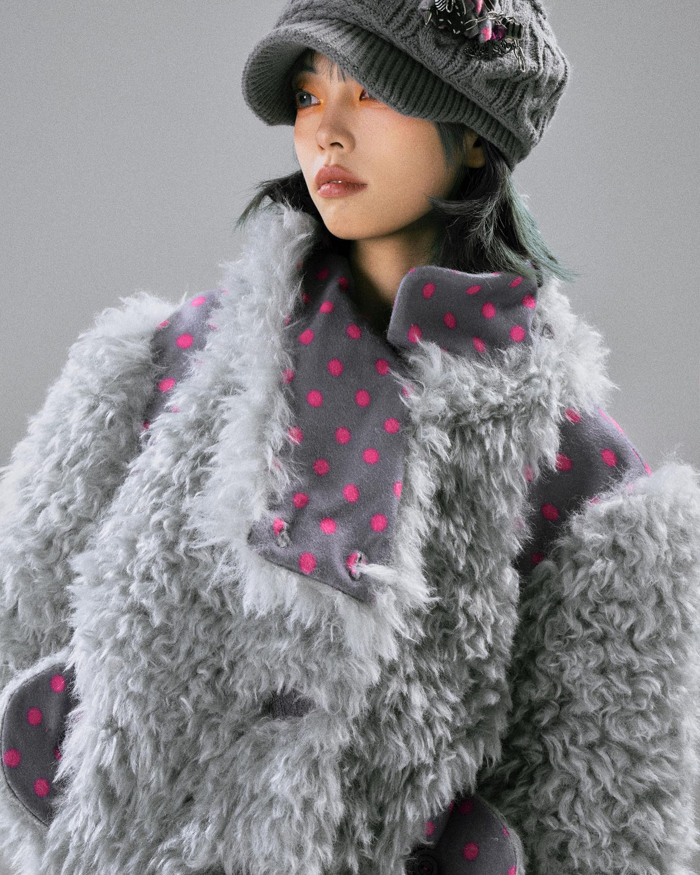 closeup shot of a woman wearing a stylish grey fluffy coat adorned with pink polka dots. the coat is textured and has a relaxed fit. she's also wearing a grey newsboy cap with embellishments. the overall style is winter fashion, and the image is professionally shot, resembling a fashion photography. the model is looking away from the camera, providing a side profile view.