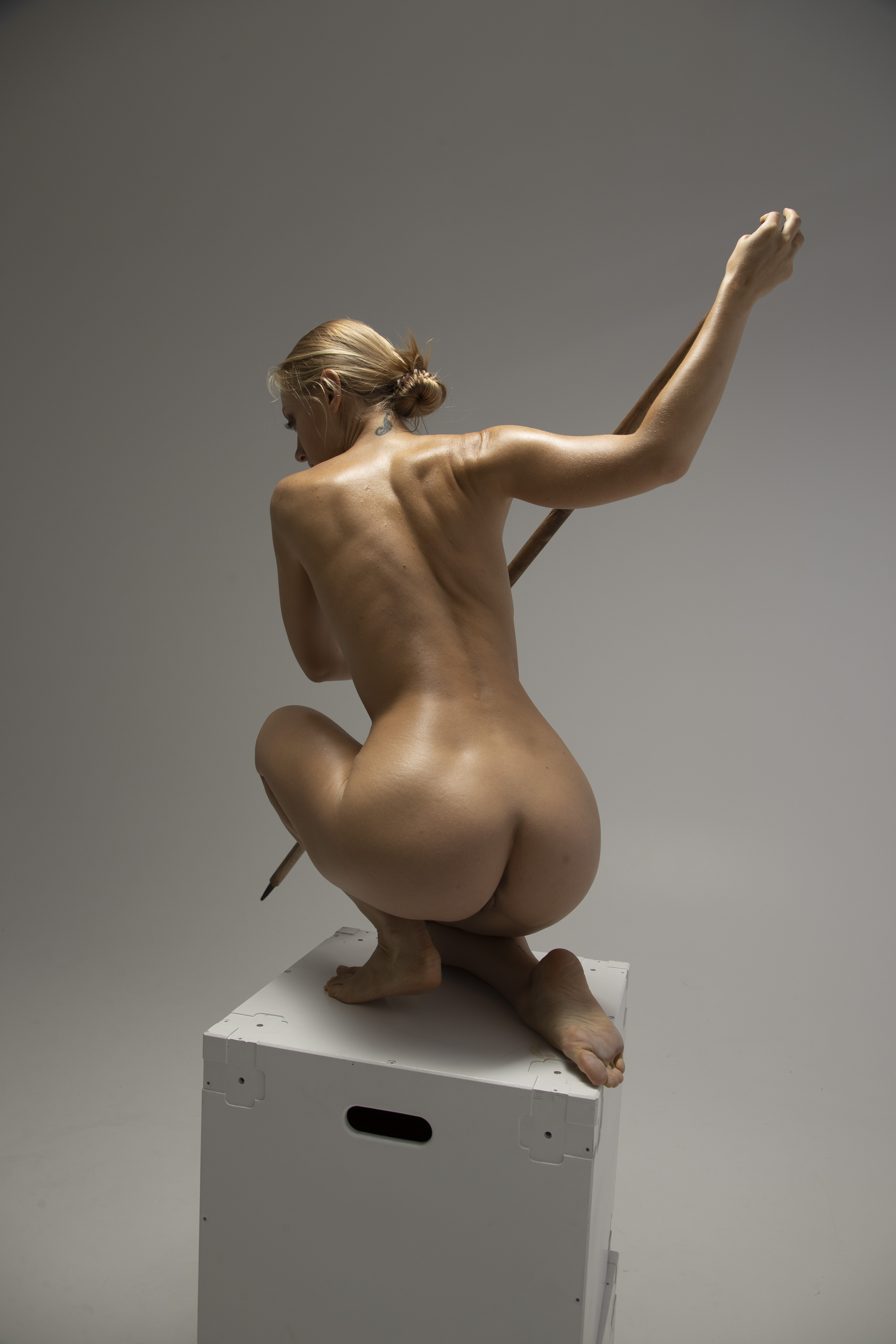 a nude woman with blonde hair pulled back in a bun is squatting on a white box. she is viewed from the back, and is holding a stick in her right hand, which is raised up. she has a tattoo on the back of her neck.