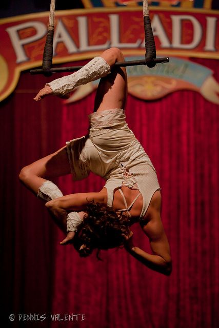 a woman dressed in a white costume performs an aerial silk routine, hanging upside down by her legs and holding onto a silk fabric. the stage is set with red curtains.