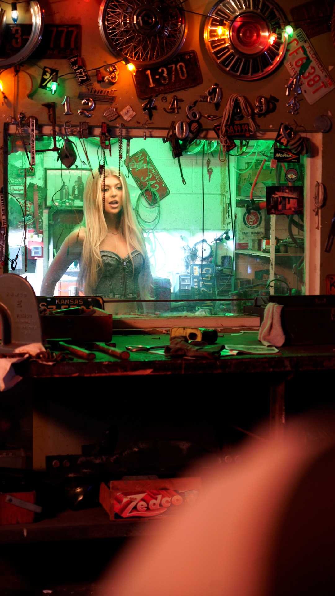 a woman with blonde hair is standing in front of a garage or auto shop window. the shop is decorated with vintage car parts and tools. the space has a green tint, and there's a zedco box visible on a shelf. the scene gives the impression of a vintage mechanic shop.