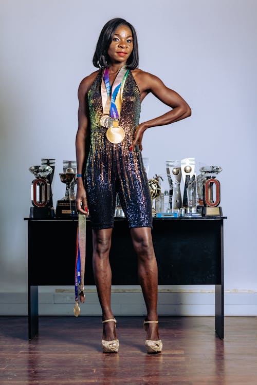 a fulllength portrait of a successful female athlete. she is standing in front of a table displaying numerous trophies and awards, wearing a shimmering black sequined jumpsuit and high heels. she confidently displays her gold and other medals around her neck. the setting appears to be indoors, against a lightgrey wall. the overall image conveys a sense of achievement and pride.