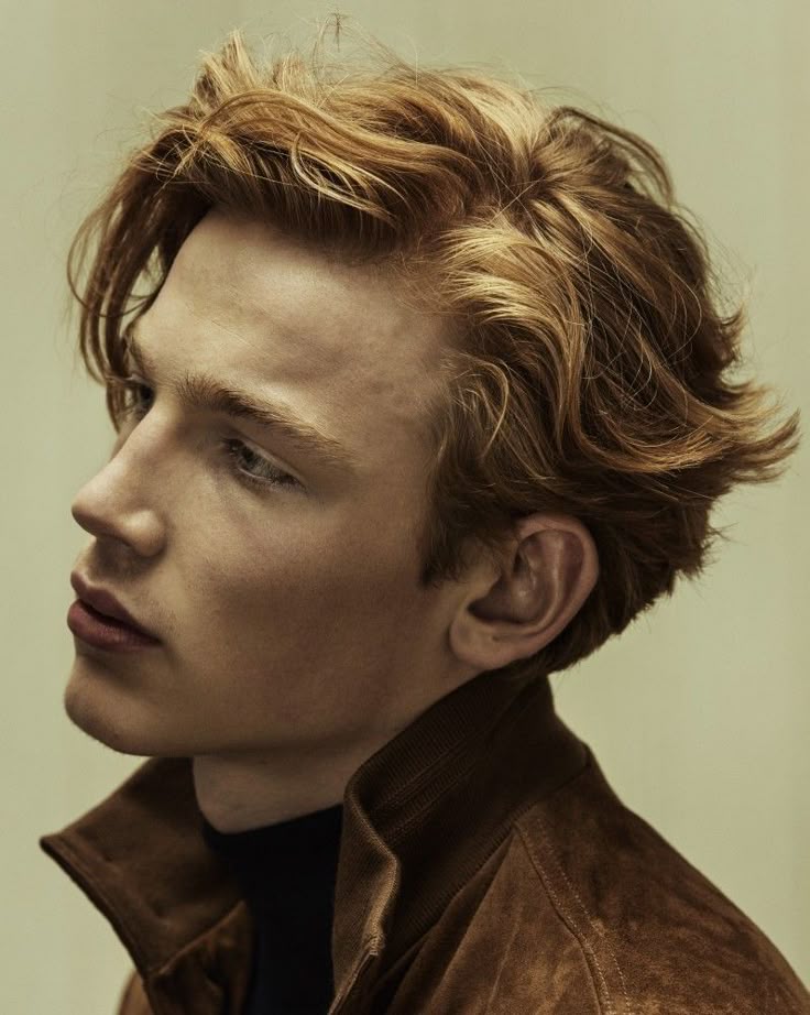 a profile headshot of a man with wavy red hair, wearing a brown suede jacket over a black turtleneck.